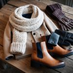 A cozy winter wardrobe laid out on a rustic wooden table, including a stylish coat, chunky knit scarf, leather gloves, and ankle boots